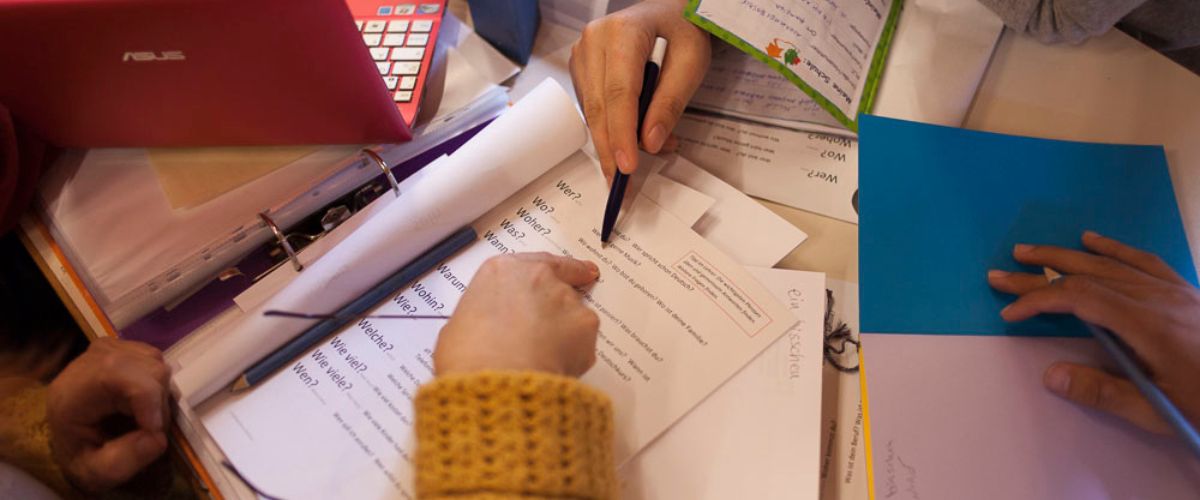 Deutsch lernen im Begegnungscafé. Hände zeigen auf Fragen auf einem Deutsch-Übungsblatt.